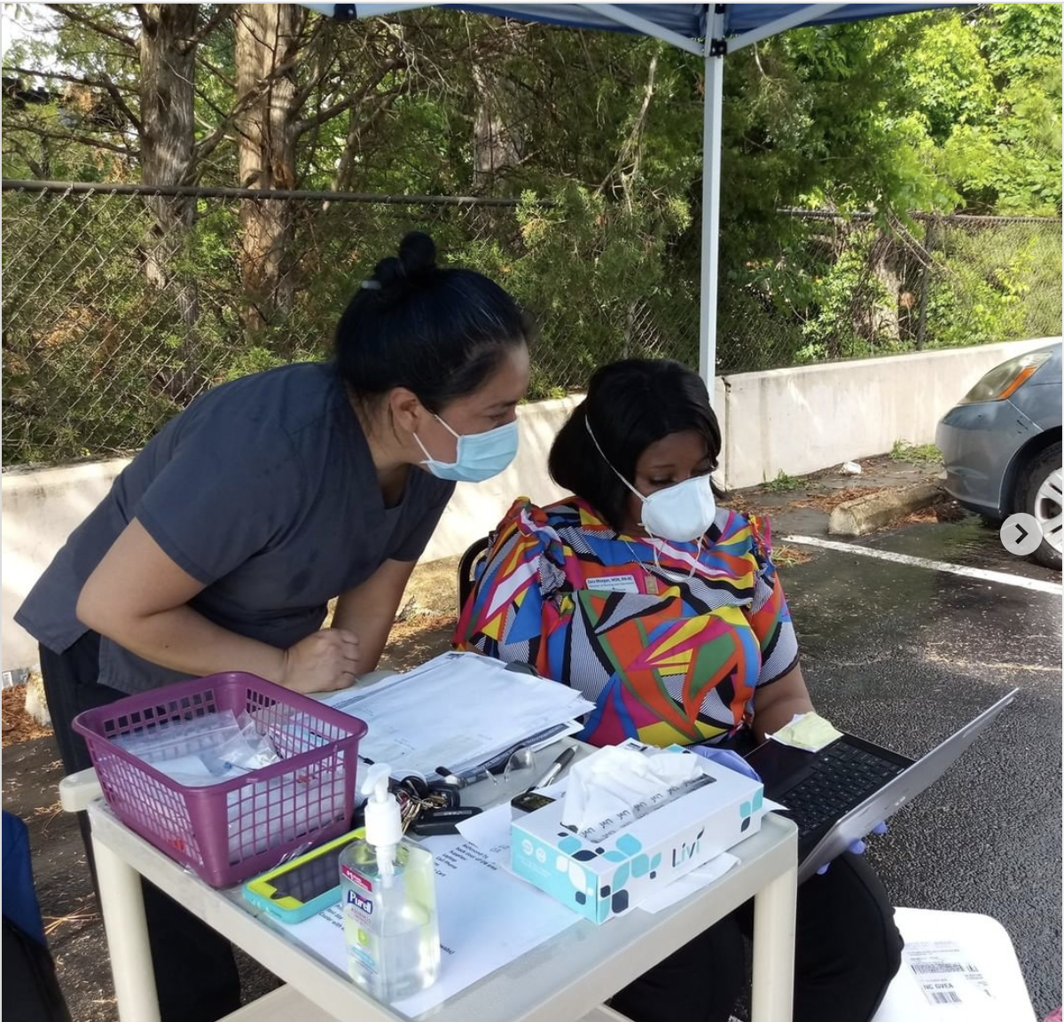 Two health care workers from CrossOver Healthcare Ministry under a tent in a parking lot, running a COVID-19 testing clinic