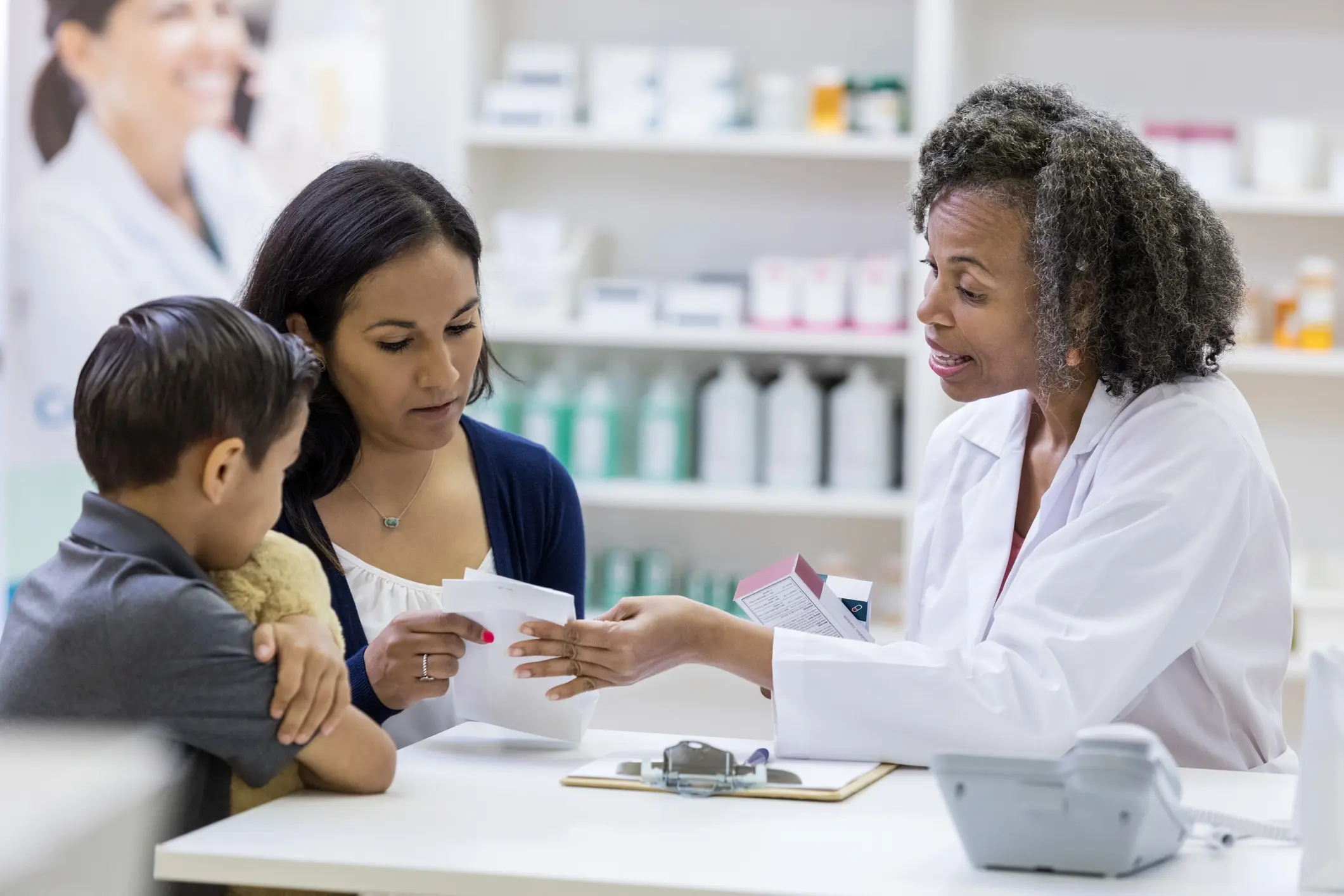 A photo of a pharmacist sitting at a table with a woman and a young boy. The pharmacist and the woman are both holding and looking at the same piece of paper while the pharmacist talks.
