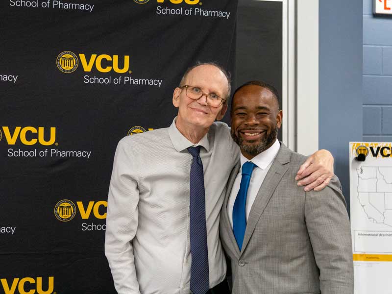 With his signature smile, Dr. David Holdford puts his arm around a smiling Dean K.C. Ogbonna in front of a VCU School of Pharmacy banner and a map of the U.S. at the Pharm.D. Class of 2026 On-Campus Day Awards Ceremony and Placement Celebration.