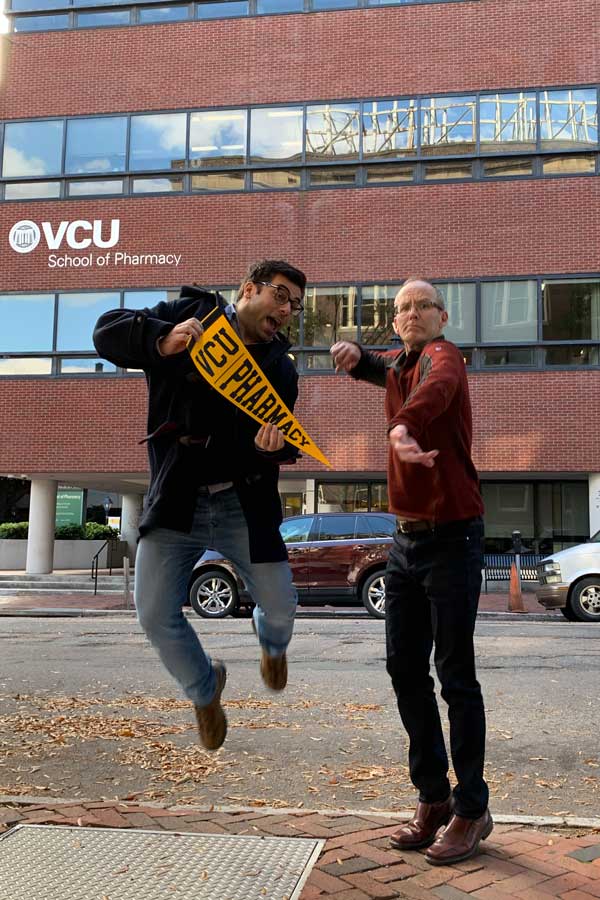 Dr. Holdford and Dr. Pontinha leap and click their heels for a photo outside VCU School of Pharmacy while Dr. Pontinha holds a VCU Pharmacy pennant just before Dr. Pontinha graduates from VCU School of Pharmacy in 2022.
