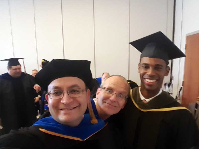 Two smiling Pharm.D. grads, Rafael Saenz and Michael Elliott, take a photo with Dr. Holdford as he makes a funny face at their graduation ceremony in 2003.