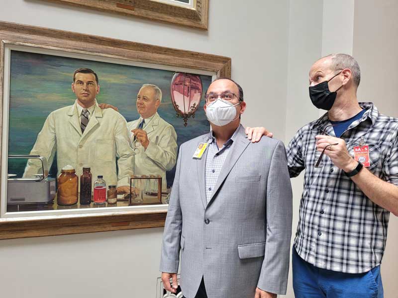 Dr. Holdford and Dr. Saenz stand next to and reenact a painting of a pharmacist mentoring a younger pharmacist with his hand on the younger pharmacist's shoulder.